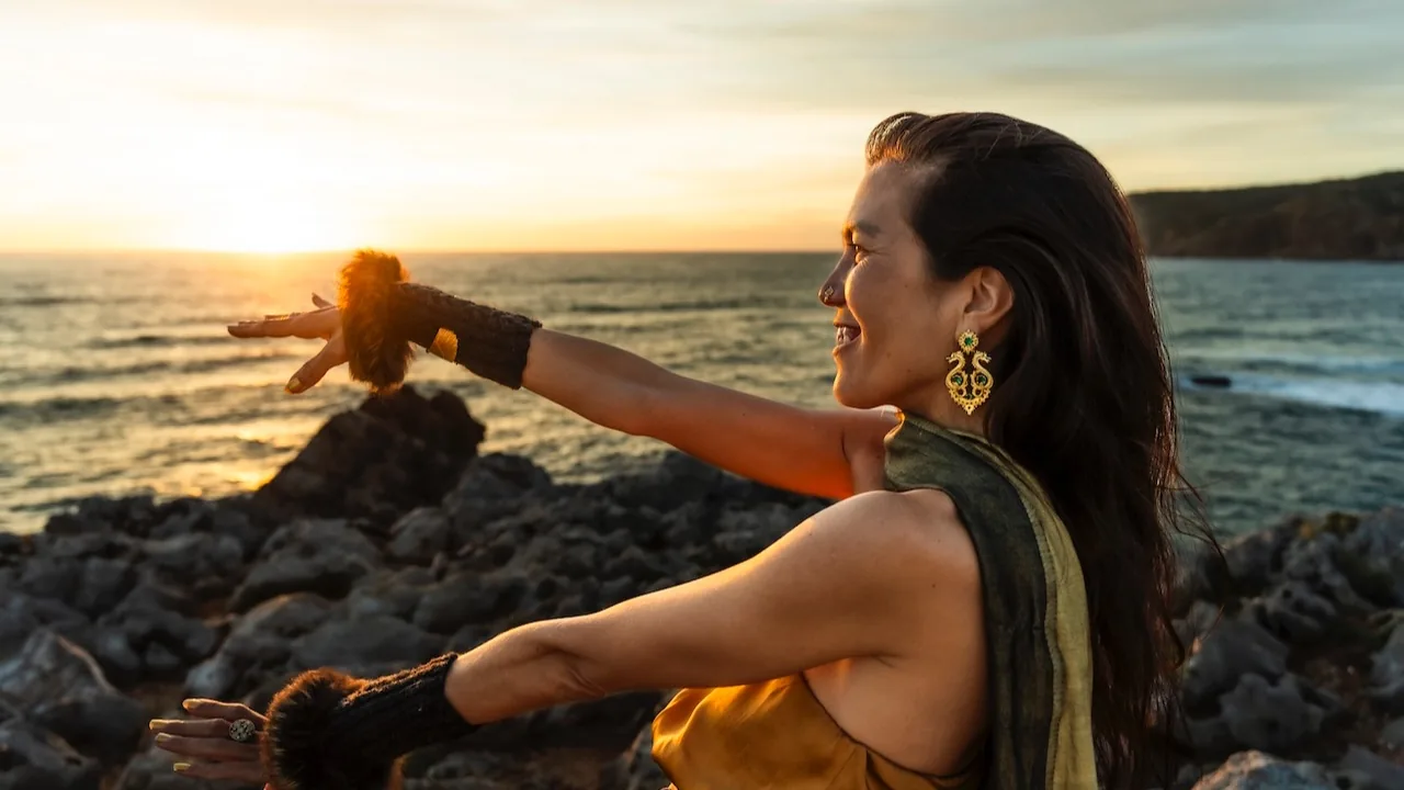 Shāradā on the rocks by the sea at sunset, smiling, one arm stretched out towards the setting sun