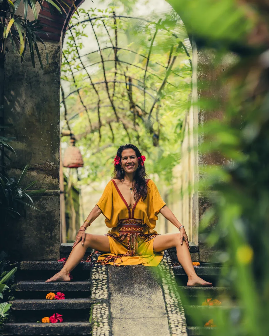 Shāradā sitting on mossy temple steps beneath a stone archway in a garden, smiling, in a saffron dress